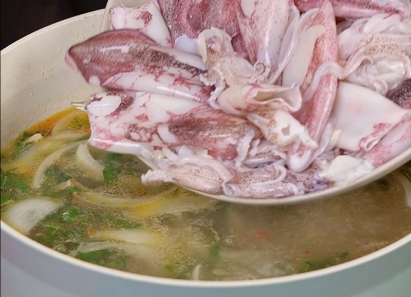 Cho mực tươi vào nồi nước dùng mẻ đang sôi cùng hành tây và các loại rau thơm